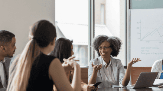 Diverse professionals in a boardroom discussion; a Black woman explains with hands raised, laptop and charts visible, J. Miller Law Firm logo in the corner.
