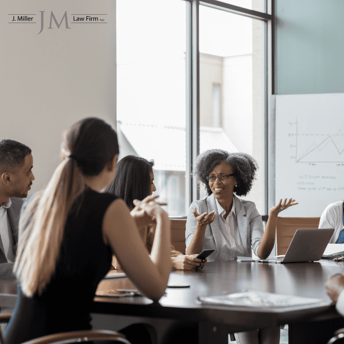 Diverse professionals in a boardroom discussion; a Black woman explains with hands raised, laptop and charts visible, J. Miller Law Firm logo in the corner.